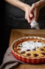 Anonyme Person in Schürze steht am Holztisch und streut Zuckerpulver auf köstlichen hausgemachten Kuchen mit Kirschen — Stockfoto