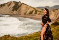 Side view of peaceful young Hispanic pregnant female in stylish dress revealing belly standing on seashore against mountains — Stock Photo