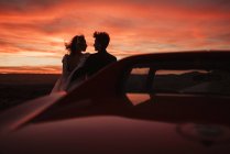 Silhouettes of bride and groom looking at each other while leaning on vehicle against cloudy sundown sky in Bardenas Reales Natural Park in Navarra, Spain — Stock Photo