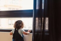 Вид сзади неузнаваемый маленький ребенок с вьющимися волосами в джинсовом комбинезоне и глядя в окно, стоя дома при дневном свете — стоковое фото