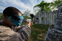 Anonymous male shooter with paintball gun aiming at enemy hiding behind wooden wall during game — Stock Photo