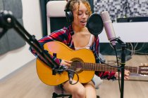 Jovem vocalista étnica feminina em fones de ouvido tocando guitarra acústica enquanto cantava com os olhos fechados no microfone no estúdio de música — Fotografia de Stock