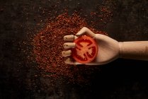 Top view composition with fresh red tomato slice in artificial wooden hand placed above ground sun dried tomatoes on black background — Stock Photo