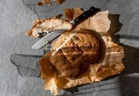 Вид зверху смачного свіжоспеченого хлібного хліба з нарізаним шматком і ножем, розміщеним на папері для випічки на сірому фоні — стокове фото