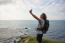 Vista laterale di escursionista maschio felice prendere auto sparato su smartphone mentre in piedi sulla collina sullo sfondo del mare durante il trekking in estate — Foto stock
