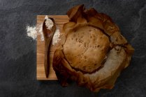 Top view composition with freshly baked rustic sourdough round bread loaf on parchment paper placed on wooden board with spoon and wheat flour — Stock Photo