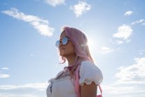 Mujer de moda con pelo rosa y gafas de sol redondas de pie sobre el fondo del mar y el cielo azul durante las vacaciones de verano - foto de stock