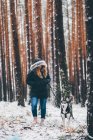 Frau in Skijacke geht mit Hund im Winterwald zwischen Bäumen spazieren — Stockfoto