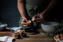 Неузнаваемый парень в фартуке разбивает сырое яйцо над миской во время приготовления теста на деревянном столе возле кухонной утвари — стоковое фото