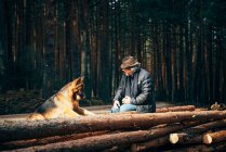 Seitenansicht des Mannes sitzt in einem Baumstamm in der Nähe von Nadelbäumen mit Haushund an sonnigen Tagen — Stockfoto