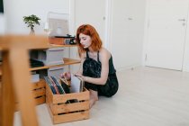 Молода жінка вибирає вініловий запис з дерев'яного контейнера, сидячи проти старовинного поворотного столу в домашній кімнаті — стокове фото