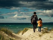 Back view of unrecognizable woman holding hand of daughter while walking together on hilly sandy seashore against wavy ocean and cloudy sky in windy weather — Stock Photo