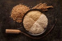 From above of bowl with assorted types of dried rice placed near bunch of ears and sickle on black background — Stock Photo