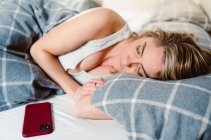 Mujer joven durmiendo en almohada suave debajo de la manta en la cama cerca del teléfono móvil por la mañana - foto de stock