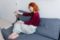 Side view of content young female sitting on sofa while talking to partner during video chat on tablet in house — Stock Photo