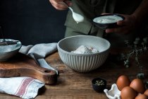 Personne méconnaissable verser du yaourt dans un bol avec de la farine près des œufs et des graines tout en préparant la pâtisserie sur une table en bois — Photo de stock