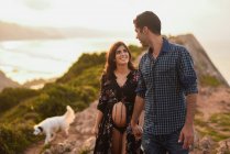 Happy Hispanic pregnant woman and loving man holding hands and looking at each other while walking with dog along hilly seashore in summer evening — Stock Photo