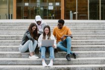 Diversi giovani amici sorridenti ampiamente mentre guardano video su netbook insieme mentre seduti su scale di cemento — Foto stock