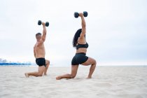Вид збоку на м'язистих багатоетнічних спортсменів, які займаються з гантелями під час тренувань на піщаному пляжі влітку — стокове фото