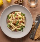 De arriba plato de pasta con rúcula, salami y nueces sobre una mesa rodeada de aceite, limón y cubiertos - foto de stock