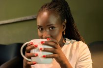 Mulher preta positiva com tranças e caneca de bebida aromática refrigerando no café e olhando para a câmera — Fotografia de Stock