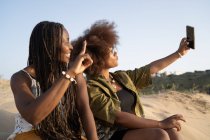 Jovens amigas afro-americanas alegres sorrindo brilhantemente enquanto tomam selfie no smartphone durante o fim de semana de verão na costa arenosa — Fotografia de Stock