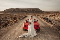 Glückliche Frau in weißem Kleid, die in der Nähe eines roten Luxusfahrzeugs steht, das auf einer staubigen Straße geparkt ist, während einer Fahrt durch den Naturpark Bardenas Reales in Navarra, Spanien — Stockfoto