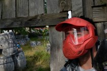 Joueur masculin masqué et avec pistolet de paintball debout derrière une clôture en bois pendant le jeu — Photo de stock