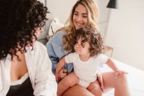Positive crop same sex couple sitting on bed and stroking little curly haired girl in bedroom — Stock Photo