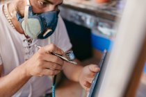 Vista lateral de artista masculino recortado en respirador usando pistola de pulverización para pintar el cuadro en lienzo durante el trabajo en taller creativo - foto de stock