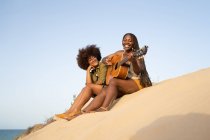Низкий угол полной длины счастливые молодые афроамериканские подруги играют на гитаре, сидя вместе на песчаном берегу моря и наслаждаясь летними каникулами — стоковое фото