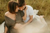 Восхитительные мужские объятия беременной женщины с закрытыми глазами, сидя на лугу в сельской местности — стоковое фото