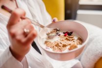 Alto ângulo de cultura fêmea irreconhecível sentado em poltrona e comer cereais saborosos com iogurte e bagas de manhã — Fotografia de Stock