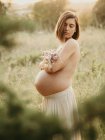 Vista laterale della donna incinta calma che copre il seno nudo con mazzo di fiori mentre si trova in campagna in estate — Foto stock