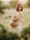 Vista lateral de la tranquila mujer embarazada que cubre el pecho desnudo con ramo de flores mientras está de pie en el campo en verano - foto de stock