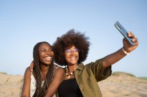 Веселые молодые афроамериканские подруги ярко улыбаются, делая селфи на смартфоне во время летних выходных на песчаном побережье — стоковое фото