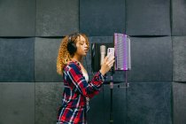 Allegro cantante afroamericana in cuffia con smartphone che canta contro scudo sonoro in studio di registrazione — Foto stock