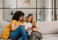 Ganzkörperfreudiges lesbisches Paar in lässiger Kleidung surft im Mobiltelefon und sitzt zusammen auf der bequemen Couch im modernen Wohnzimmer — Stockfoto