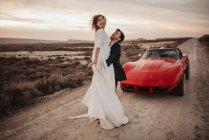 Full body groom hugging and lifting bride while standing on road near red luxury car in evening in Bardenas Reales Natural Park in Navarra, Spain — Stock Photo