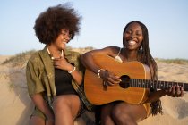Felices amigas afroamericanas jóvenes tocando la guitarra mientras están sentadas con los ojos cerrados en la orilla del mar y disfrutando de las vacaciones de verano - foto de stock