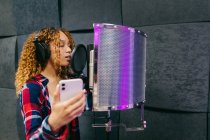 Allegro cantante afroamericana in cuffia con smartphone che canta contro scudo sonoro in studio di registrazione — Foto stock