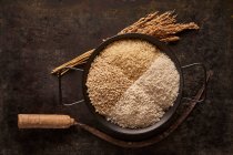 From above of bowl with assorted types of dried rice placed near bunch of ears and sickle on black background — Stock Photo