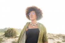 Мечтательная молодая афроамериканка с вьющимися волосами, одетая в стильную летнюю одежду с ожерельем и очками, отворачивающаяся, стоя в солнечном свете на берегу моря летом — стоковое фото