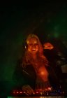 Feliz joven DJ femenino en auriculares sonriendo y utilizando sintetizador para reproducir música mientras está de pie en el humo durante la fiesta en el club nocturno - foto de stock