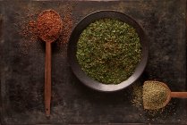 Top view of bowl with green dried herbs and spoon with ground sun dried tomato powder with artificial wooden hand on black background — Stock Photo