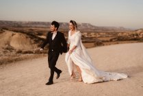 Allegro sposo e sposa che cammina tenendosi per mano vicino alla montagna contro il cielo nuvoloso al tramonto nel Parco Naturale di Bardenas Reales in Navarra, Spagna — Foto stock