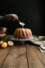 Vista de la cosecha de recubrimiento manual con una cuchara un pastel de esponja de limón colocado en una mesa de madera sobre un fondo oscuro - foto de stock