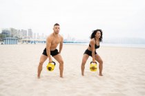 Multiracial fit sportsman and sportswoman doing kettlebell swing exercise while training at seaside in summer — Stock Photo