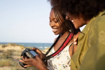 Vista lateral de alegres jóvenes amigas afroamericanas con ropa de verano y accesorios elegantes revisando fotos en la cámara fotográfica mientras pasan las vacaciones de verano juntas en el campo - foto de stock