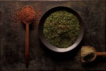 Top view of bowl with green dried herbs and spoon with ground sun dried tomato powder with artificial wooden hand on black background — Stock Photo
